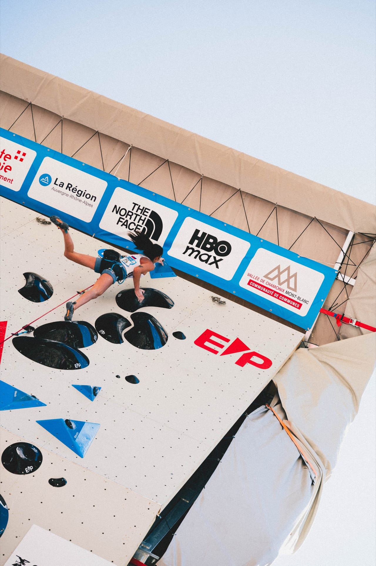 Low-angle shot looking up at a climber on the IFSC wall, sponsors banners visible